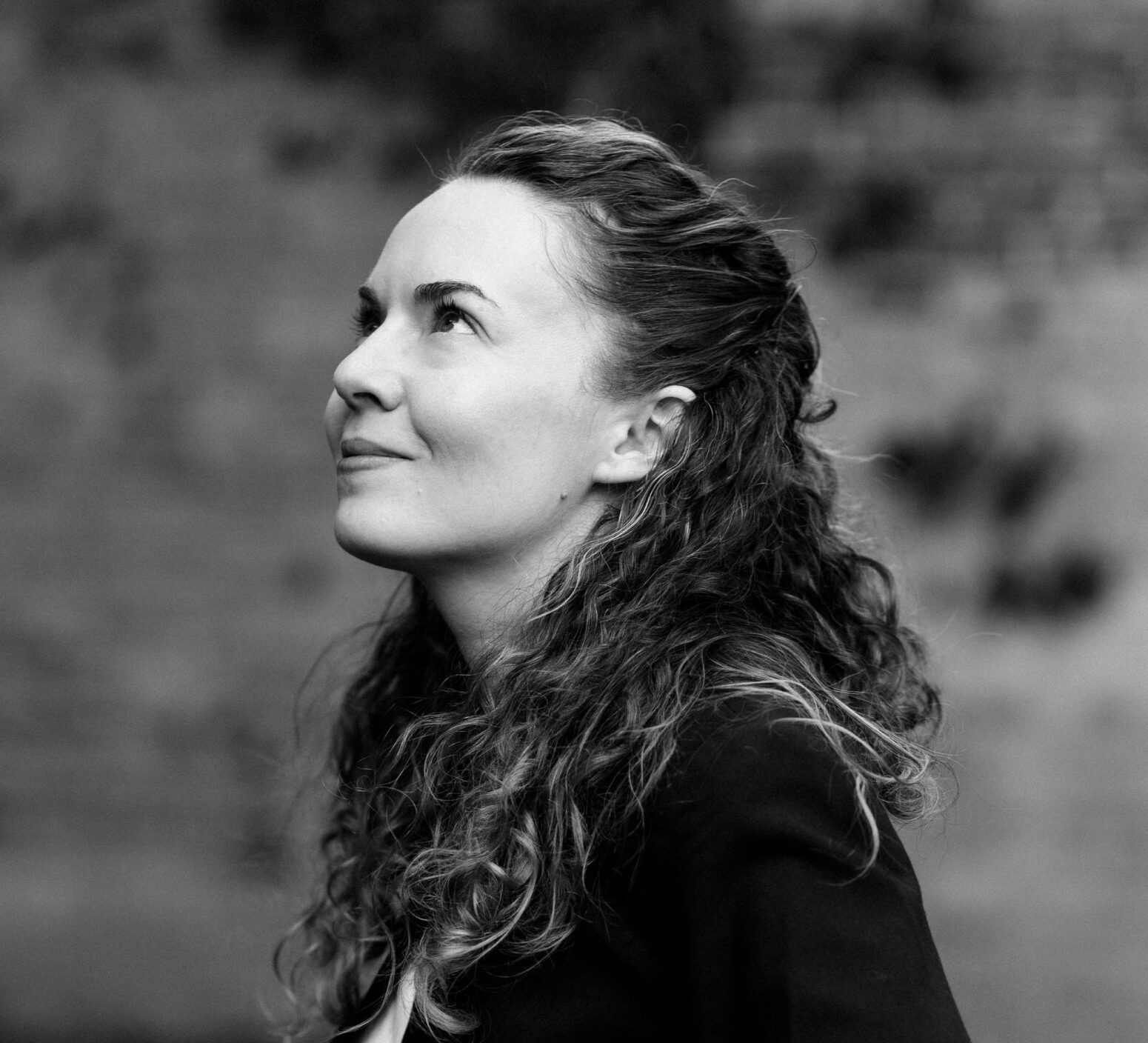Bess Winter Kosinec, a white woman with long curly hair, wears a black blazer and white tee and stands in profile, looking up and smiling. Photo is black-and-white.