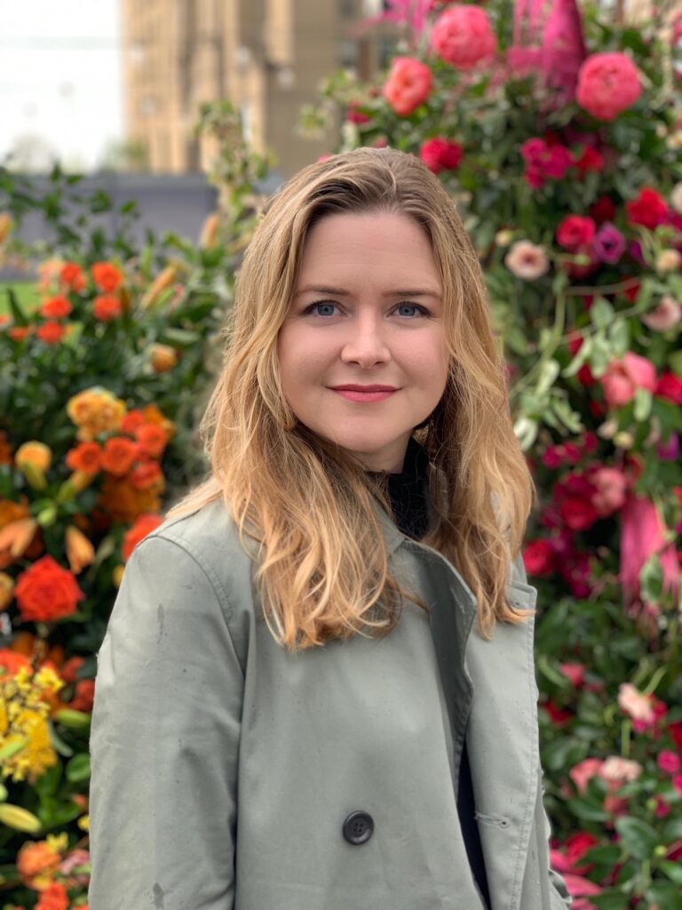 Kate McIntyre, a white woman with wavy blond hair, stands in front of a trellis of bright flowers. She is wearing a gray coat.