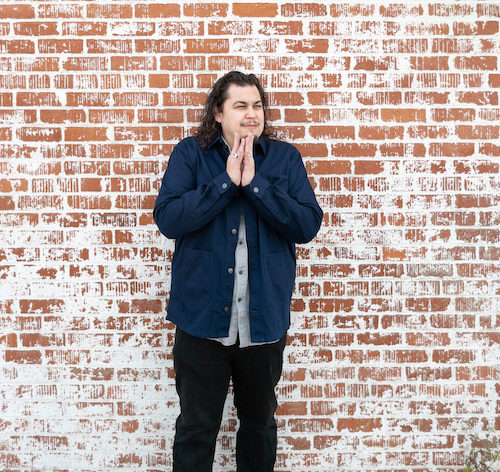 Anthony Cody, a Latinx poet, stands before a red brick wall. He wears a denim shirt and black pants and holds his hands clasped to his chest. He looks away from the camera. 