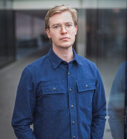 A Ukrainian-American man with short, blonde hair. He stands in front of a city building, wearing a blue button-down and round-shaped glasses.