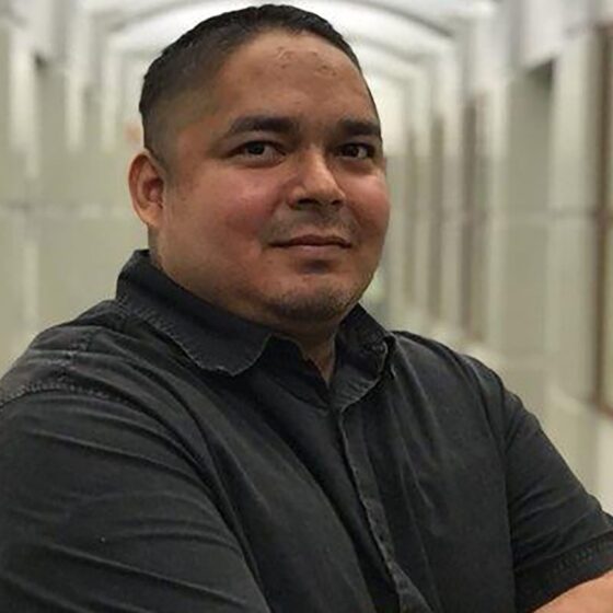 A Latino man in a black short-sleeved, collared shirt. He's standing in a hallway.