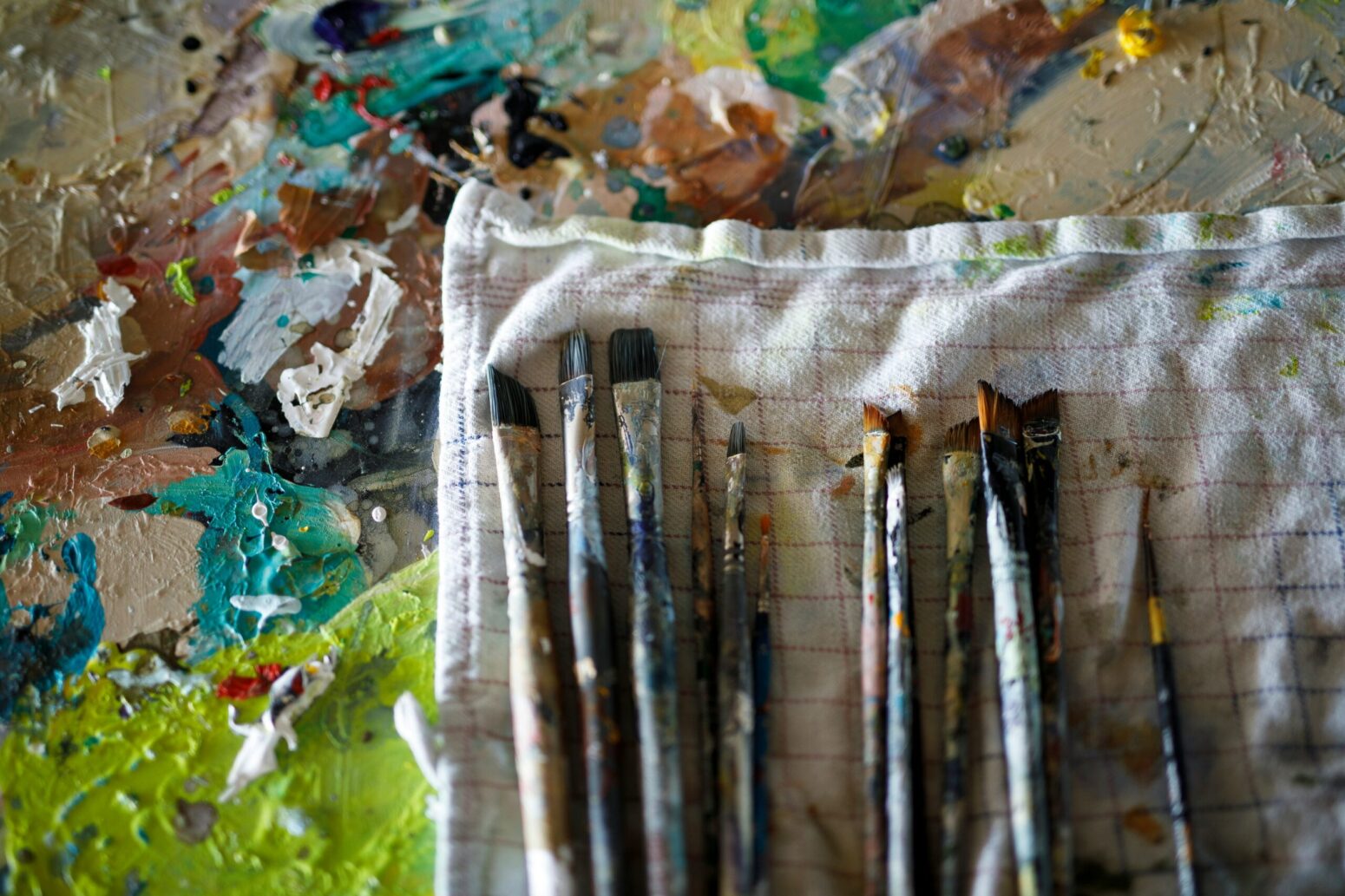 A bunch of small, dirty paintbrushes sitting on a white cloth. The cloth rests on a table, covered in splatters of paint.