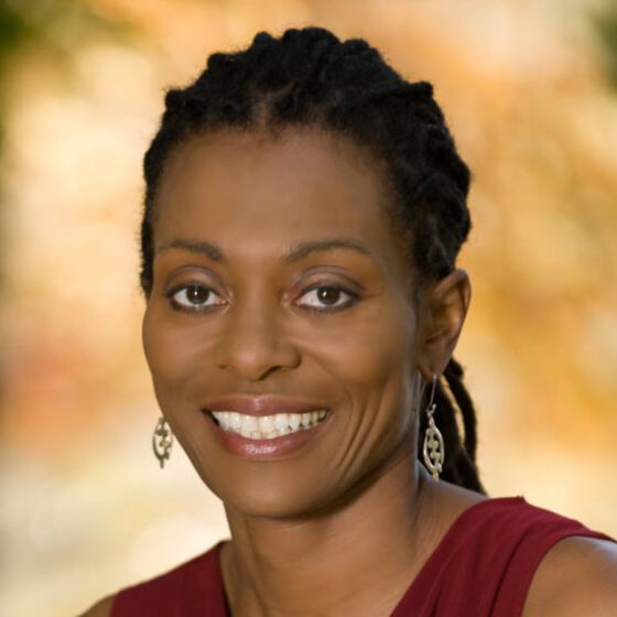 A Black woman with braids, dangling earrings, and a maroon sleeveless top. She is smiling with blurred greenery behind her.