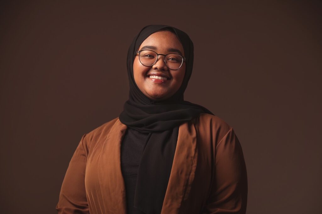 Fatma Omar, a young Black woman wearing round tortoiseshell glasses, a black hijab, and a brown jacket, smiles at the camera in front of a dark-brown backdrop.