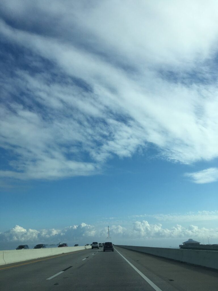 View of the Sunshine Skyway Bridge looking forward out of a car windshield. There are cars in the lanes ahead. The bridge, itself, only takes up the lower fifth of the photo; the rest is blue sky with swaths of white clouds.