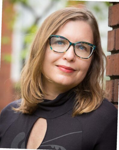 Jenny Bitner, a white woman with light brown hair, glasses with a bluish frame, and a black keyhole-style shirt, stands outside next to a brick building with a tree and another building's edge blurred behind.