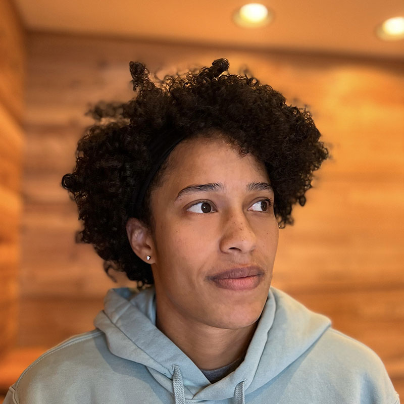 Parrissa Eyorokon, a Middle Eastern/North African woman with short curly hair, stands in front of a warm wood background. She looks to the side, and wears a gold earring and gray hoodie.