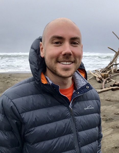 Matt Barrett, a white man in a black puffy Eddie Bauer coat slightly unzipped and orange shirt, stands smiling on a beach with gray sky, waves, sand, and a pile of driftwood behind him.