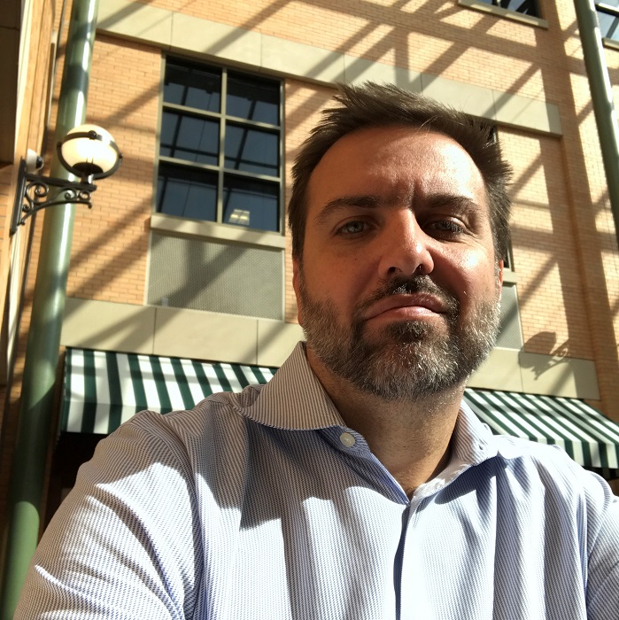 Amorak Huey, a white man with dark hair and a salt-and-pepper beard. He's wearing a blue-and-white vertically striped shirt, and he's in sunshine with a building and awning behind him.