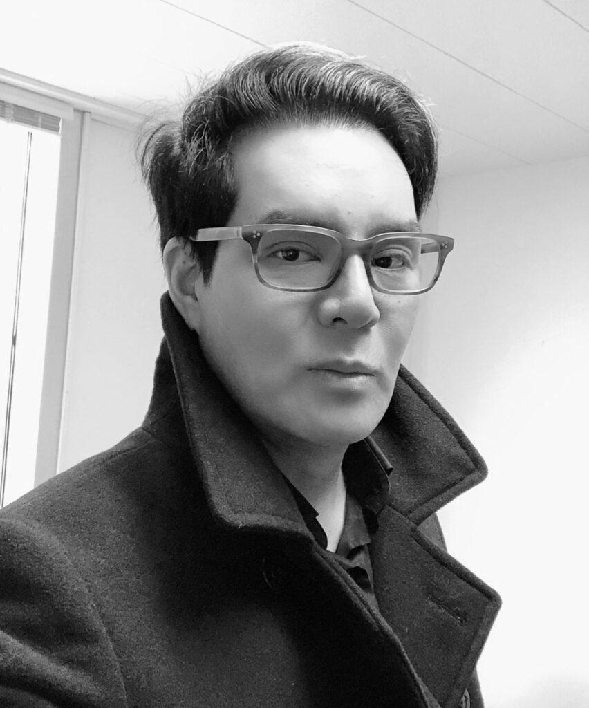 Kenneth Tanemura, an Asian-American man, looks at the camera in a black-and-white shot. He wears a coat with a turned-up collar, and square glasses. He appears to be inside; there is a window behind his shoulder.