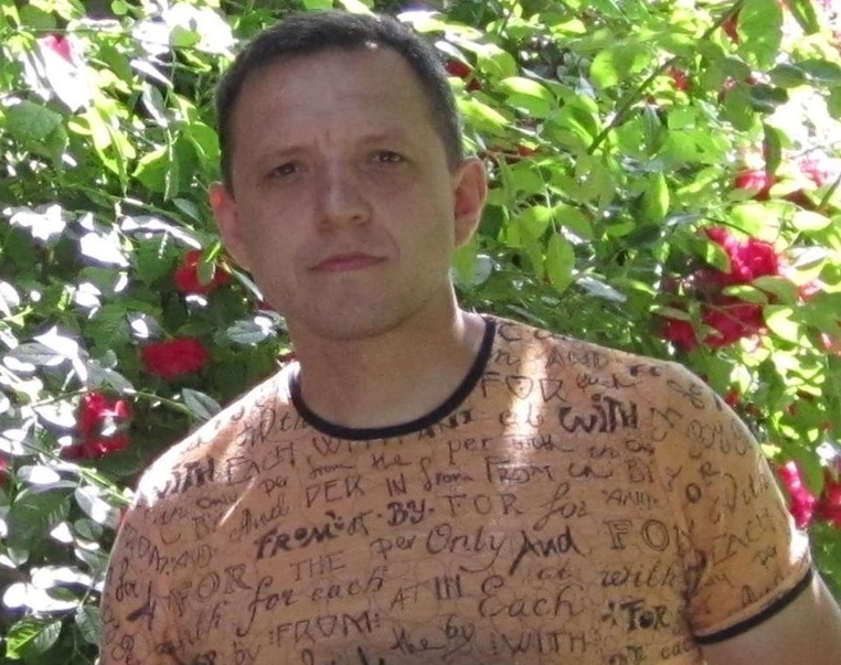 Dmitry Blizniuk, a Ukranian man in a light orange shirt with prepositions written on it and a black crewneck collar, stands in front of a bush with flowers. 