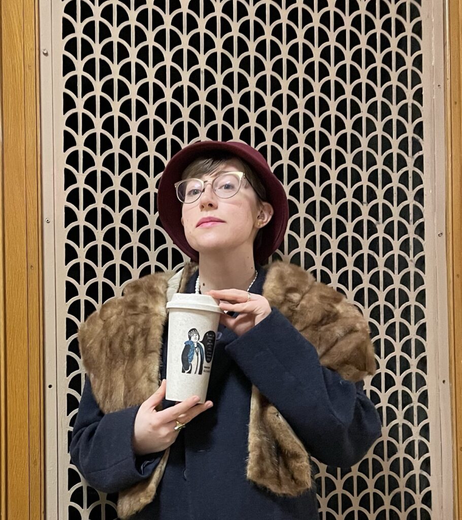 Assistant Editor Lily Davenport dressed in a circa-1920s George Elliston costume, with maroon cloche, blue coat, brown mink stole, and long string of pearls. She stands in front of a deco-style grate and proudly holds the CR eco-mug in front of her as she looks down at the camera.