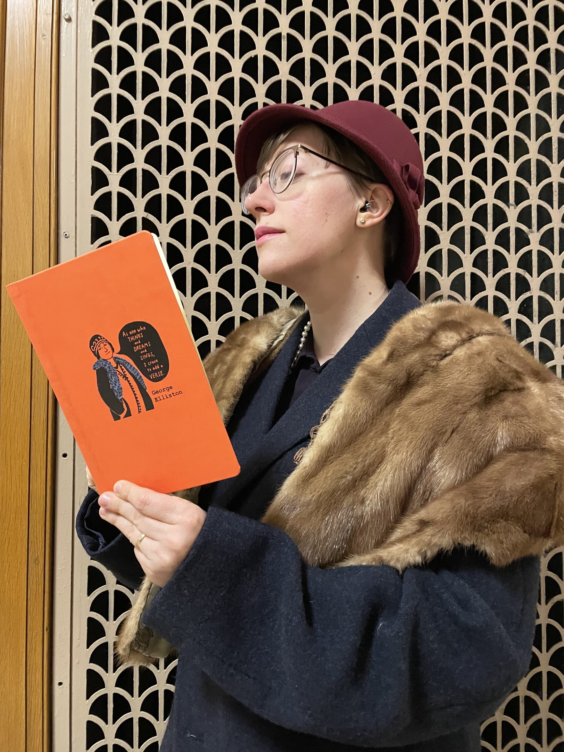 Assistant Editor Lily Davenport dressed in a circa-1920s George Elliston costume, with maroon cloche, blue coat, brown mink stole, and long string of pearls. She stands in profile in front of a deco-style grate and reads from CR's perfect-bound orange notebook.