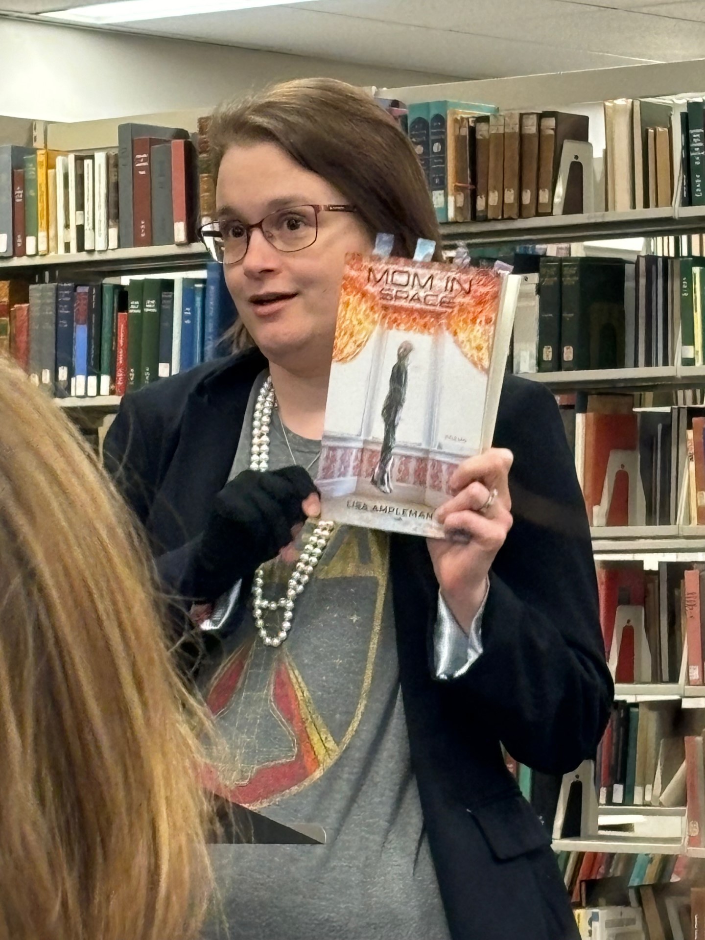 Lisa Ampleman, a white, brown-haired woman in glasses, blazer, NASA tee shirt, and string of pearls, holds her book, MOM IN SPACE, up as she speaks to the audience at Poetry Stacked. She stands in front of a large bookcase filled with books.