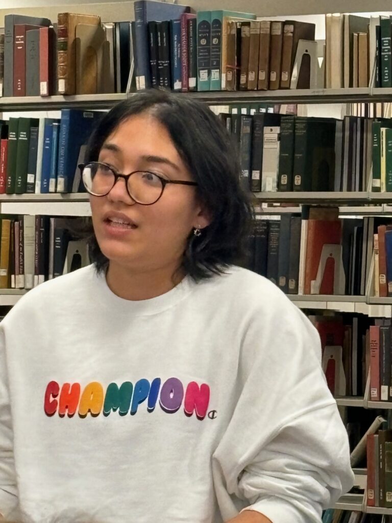 Dani Charles, a young Latinx poet wearing glasses and a white Champion sweatshirt, reads in front of a bookcase filled with books. 
