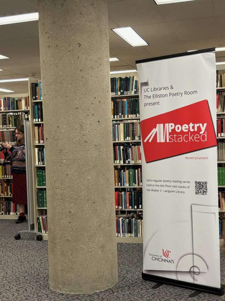 A vertical banner advertising the Poetry Stacked series hangs from a stand in the stacks, next to a large concrete column. A woman stands to the left of the column, partly obscured by a bookshelf.