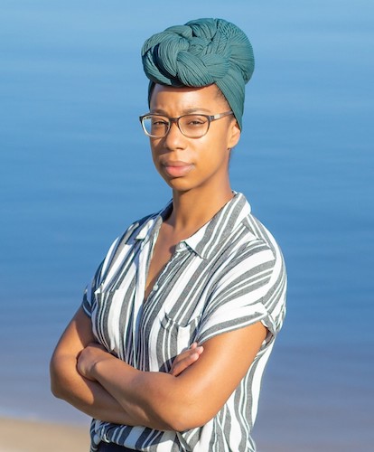 Chet'la Sebree, a Black woman wearing a grey and white striped shirt, rectangular eyeglasses, and a light blue headwrap, looks into the camera in front of a piercing blue ocean background. 