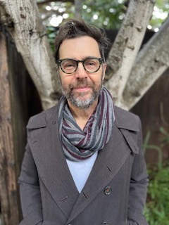 Ryan Griffith, a middle-aged white man with brown hair and salt-and-pepper beard and mustache, stands in front of a tree. He wears a gray overcoat and striped infinity scarf, and gray tortoiseshell glasses, and smiles.