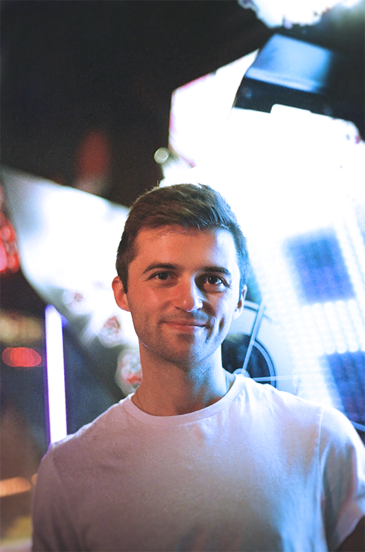 Michael Colbert, a clean-shaven white man with short brown hair, smiles at the camera. He wears a white tee shirt and is standing against a brightly lit nighttime background, with neon lights.