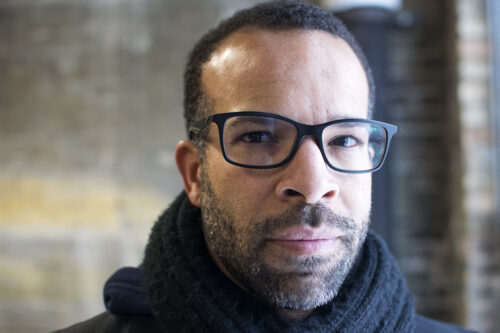 Douglas Kearney, a Black man with a beard and glasses, wearing a thick black scarf. The background behind him, a brick wall, is blurred.