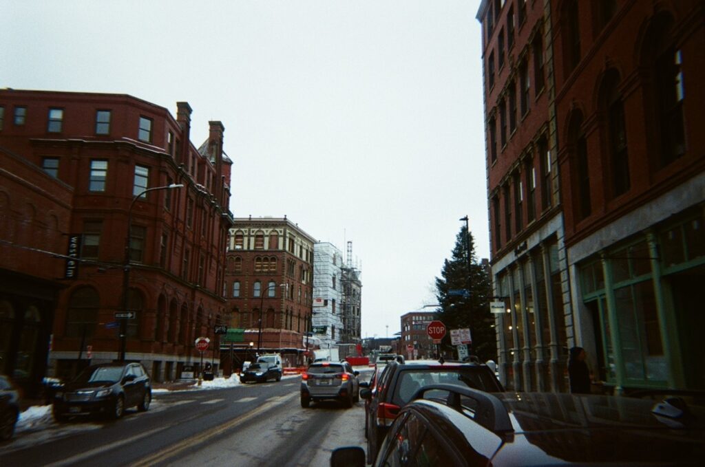 A city streetscape, taken from beside a parked car. It's winter; the sky is gray and there's snow on the ground. Cars pass by. Most of the buildings are red brick.