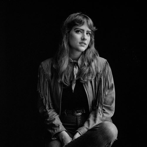 A black and white photo of the poet, Rebecca Griswold. She stands before a solid black background and looks into the camera, wearing a western-style leather jacket with fringe and blue jeans. She has long brown/blonde hair and wears a nose ring. 