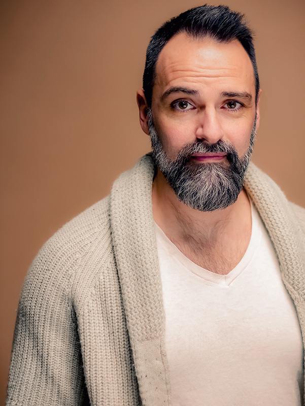 Sean McIntyre, a white man with dark cropped hair and salt-and-pepper beard, stands in front of a tan backdrop. He wears a cream v-neck tee under a tan shawl-collared sweater.