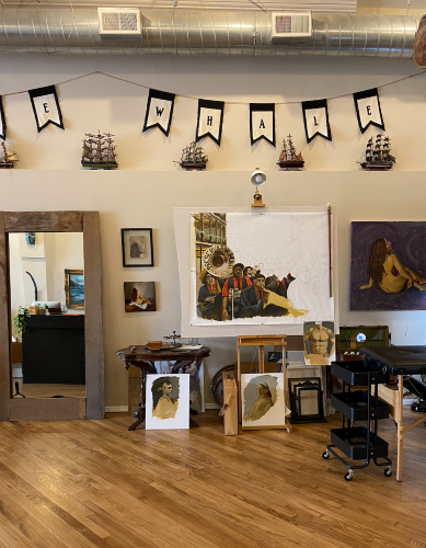 The interior of White Whale Tattoo and Gallery. Oil paintings in various states of completion hang on the wall and rest against easels. A "White Whale" banner hangs from the ceiling, and a series of model-ships are displayed prominently below it. 