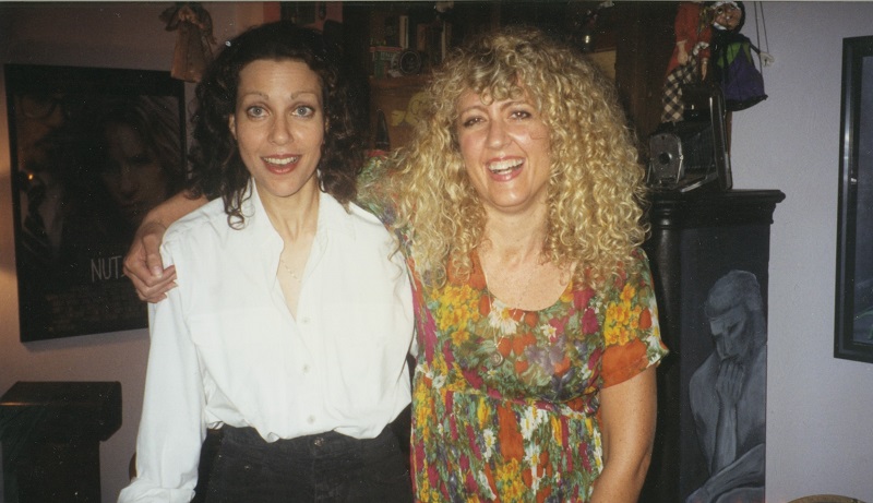 A snapshot of two white women. On the left is a brunette with a smile and slightly surprised expression, wearing a white button-down shirt and dark pants. The other woman, on the right, has her arm around the brunette. The latter has curly blond hair and a large smile, and she's wearing a multicolored flower empress-waist dress.
