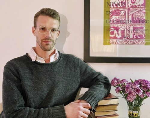 Daniel Uncapher, a white man with blond hair, a closely cut beard, and white wire-rimmed classes, sits beside a colorful woodblock print and a vase of pink flowers. He wears a green knit sweater over a white collared shirt.