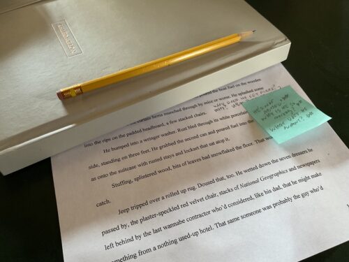 A white bound notebook and pencil on top of a printed draft of a story, with a blue post-it note that has questions in pencil on it.