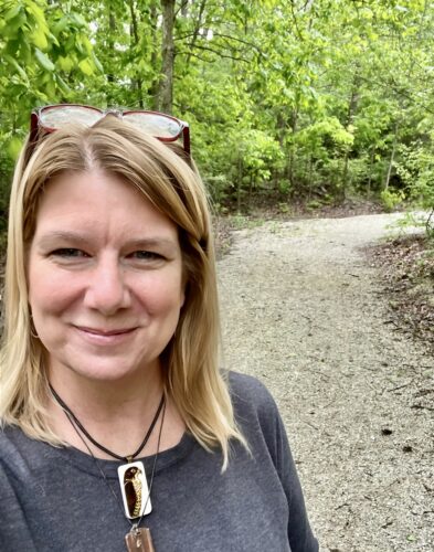 Jolene McIlwain, a white woman with blond hair and a pair of glasses on the top of her head, stands on a gravel path in a wooded area. She's wearing two necklaces, one with a hawk on it, and she's smiling.