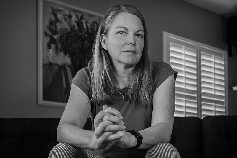 Black-and-white shot of Michelle Ross, a white woman with chest-length blond hair, sitting on a couch with her hands clasped. She looks slightly sideways. She wears a tee and small pearl necklace. Behind her, a painting of flowers and a shuttered window.