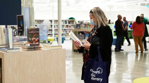 A woman with blond hair, a black headband, and a mask holds a black Cincinnati Public Library bag as she looks at a book. A set of shelves with some books on top on display is to the left.