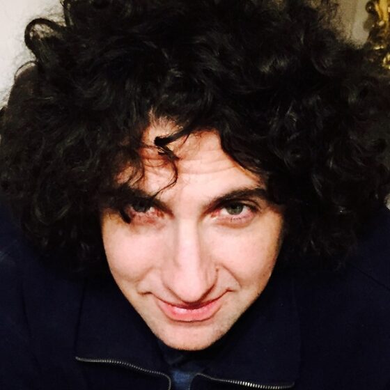 A head shot of a man with large curly black hair. He's leaned forward, smiling slightly, and wearing a dark jacket