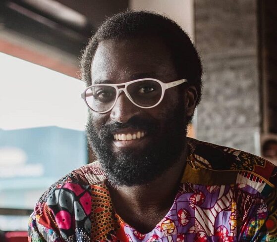 An African person with dark hair and a full beard, smiling. They are on a porch outside and wearing white-framed glasses and a multicolored shirt.