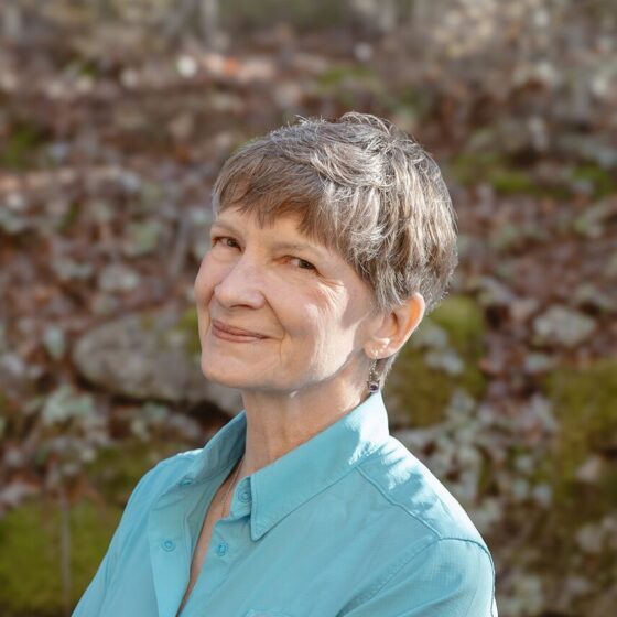 A white woman with gray hair in a pixie cut, smiling facing the left. She is outside near a forest, and wearing a cyan button-down.