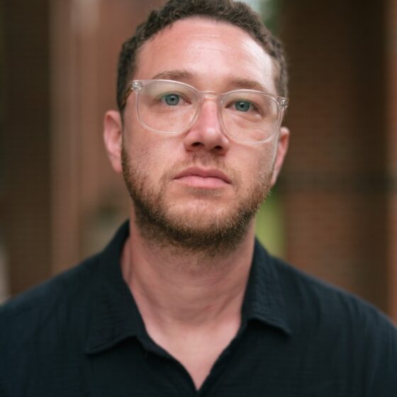 A multiracial man with brown, curly hair and a slight beard. He stands in front of a blurred, brown-and-white background, and wears a black button-up and clear, square-shaped glasses.