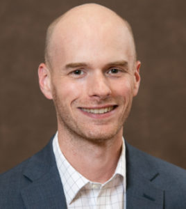 A bald, white man, smiling. He is standing in front of a brown background and wearing a white button-up with a charcoal suit jacket overtop.