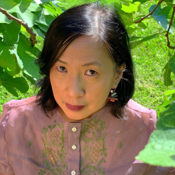 An Asian-American woman with shoulder-length, straight, black hair, smiling. She is standing outside and wearing a pink button-up with green detailing.