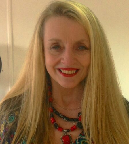 A white woman with long blond hair, blue eyes, a smile with red lipstick, and a black and red necklace.