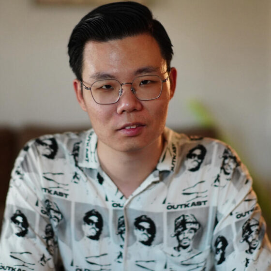 An Asian man with short, black hair. He is sitting in a white-walled room, and wearing a black-and-white button-up with peoples' pictures on it, and square-shaped glasses.