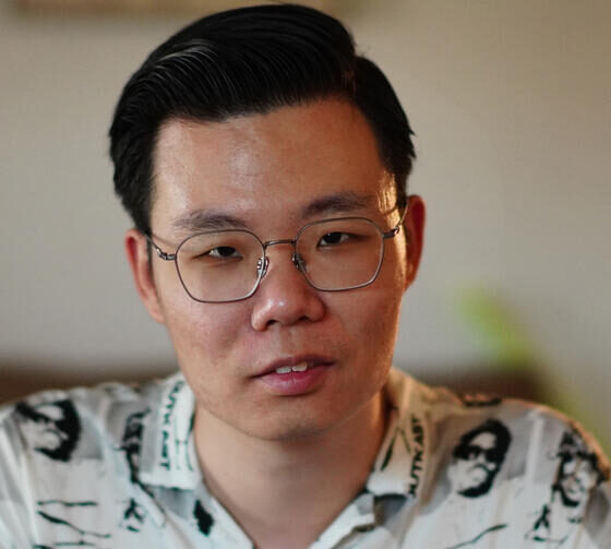 An Asian man with short, black hair. He is sitting in a white-walled room, and wearing a black-and-white button-up with peoples' pictures on it, and square-shaped glasses.