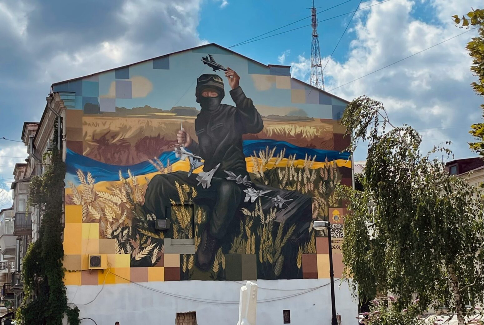 A photo of a mural, known as the "Kyiv Avenger" in Kyiv, Ukraine. It is a painting of a Ukrainian soldier in full gear, sitting in a field of grain with a long, Ukrainian flag laid out behind them. The soldier weaves together small, battle airships.