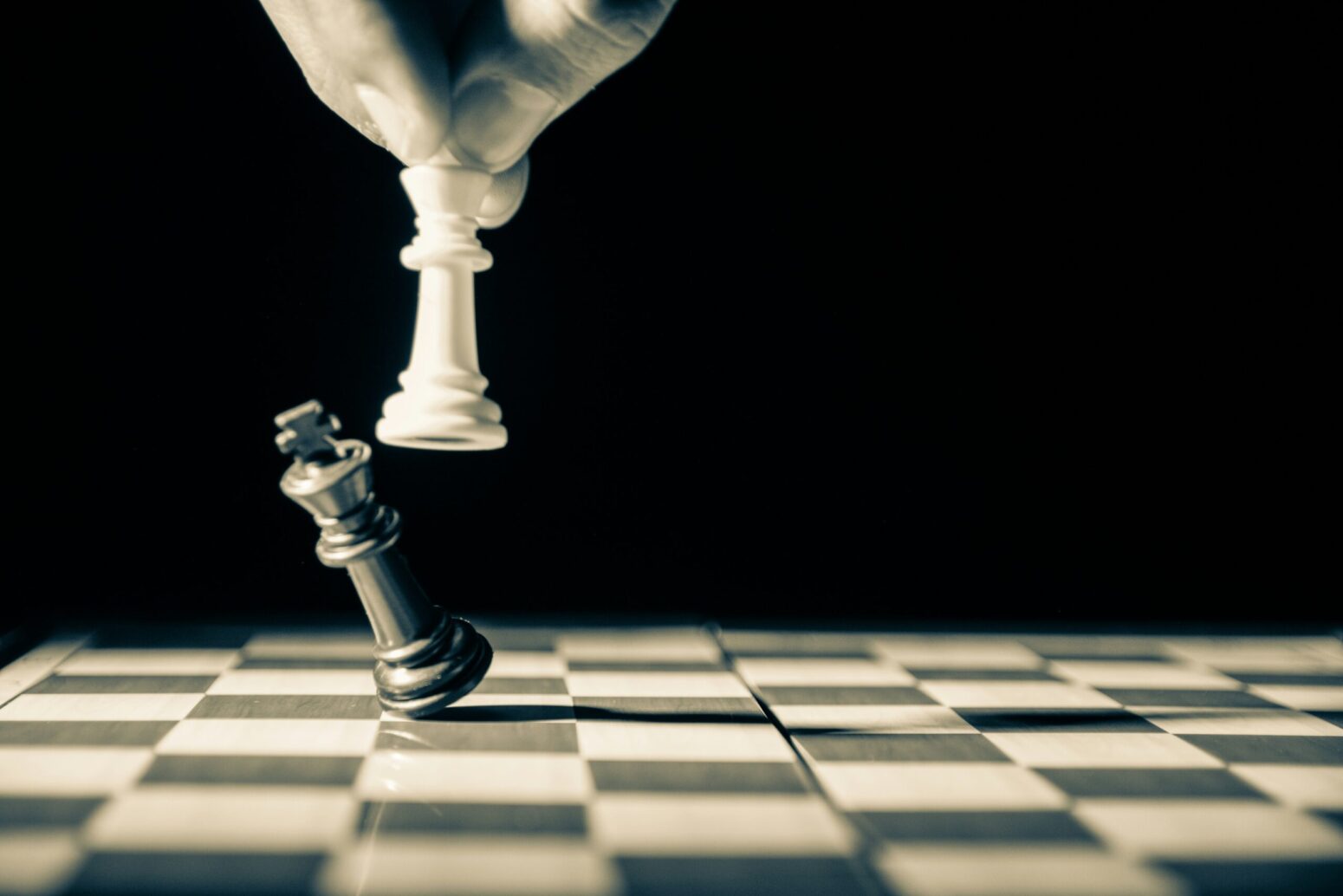 A black-and-white photo of a chess board with only two pieces on it. The black king is being knocked down by the white king by someone's hand.
