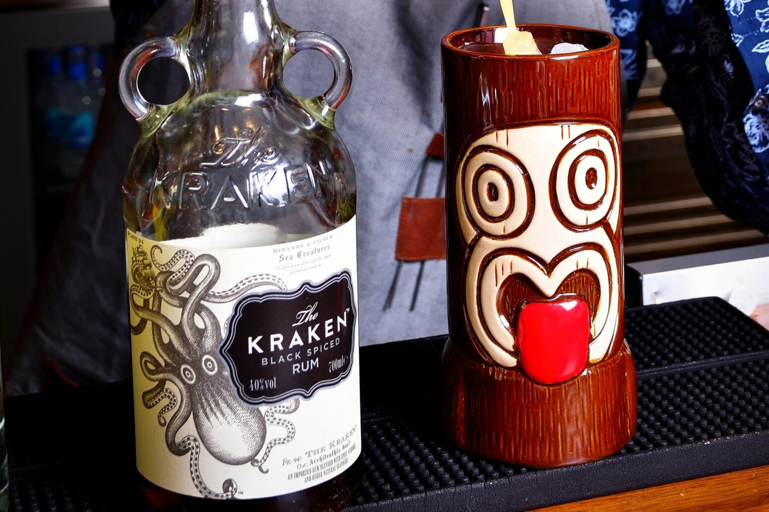 A jug of a drink called "Kraken: black spiced rum" sits on a bar. It is to the left of a cup that has been painted to look like a tiki head with its tongue sticking out. Someone behind the bar wearing a gray apron is pouring a creamy liquid into the tiki cup.
