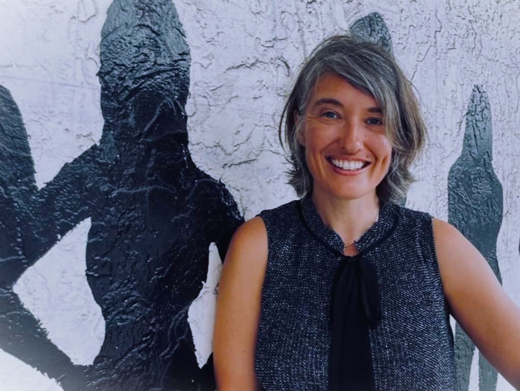 A white woman with gray shoulder-length hair, smiling and wearing a gray sleeveless blouse with black ribbon. She's standing next to a shadow painted on a wall.