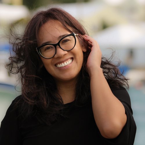 A multiracial woman with wind-swept, brown hair that goes past shoulders, smiling and tilting her head to the left. She is tucking her hair being her right ear. She's outside near a harbor, and wearing a black shirt and cat-eye-shaped glasses.