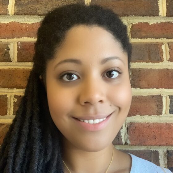 Erica Dawson, a Black woman with long braids over her right shoulder, with a gold necklace that says E and a light-blue shirt. She is smiling and standing in front of a red-brick wall