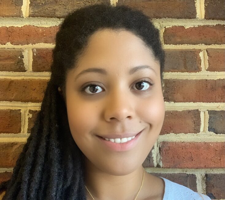 Erica Dawson, a Black woman with long braids over her right shoulder, with a gold necklace that says E and a light-blue shirt. She is smiling and standing in front of a red-brick wall